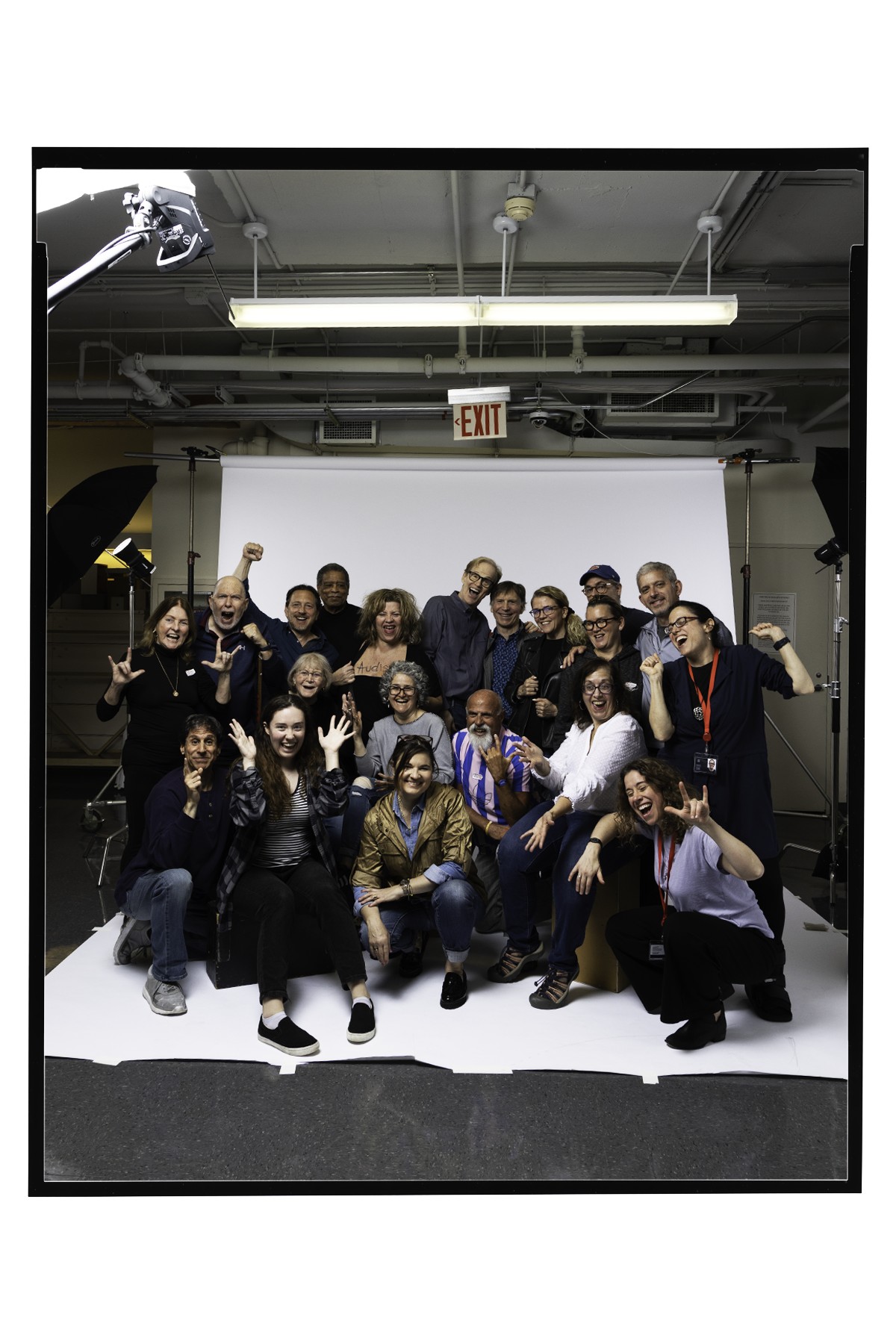 A large group of people pose together – some are signing “I love you” or are cheering in ASL. The group is on a large white backdrop with lighting and camera equipment in the background.