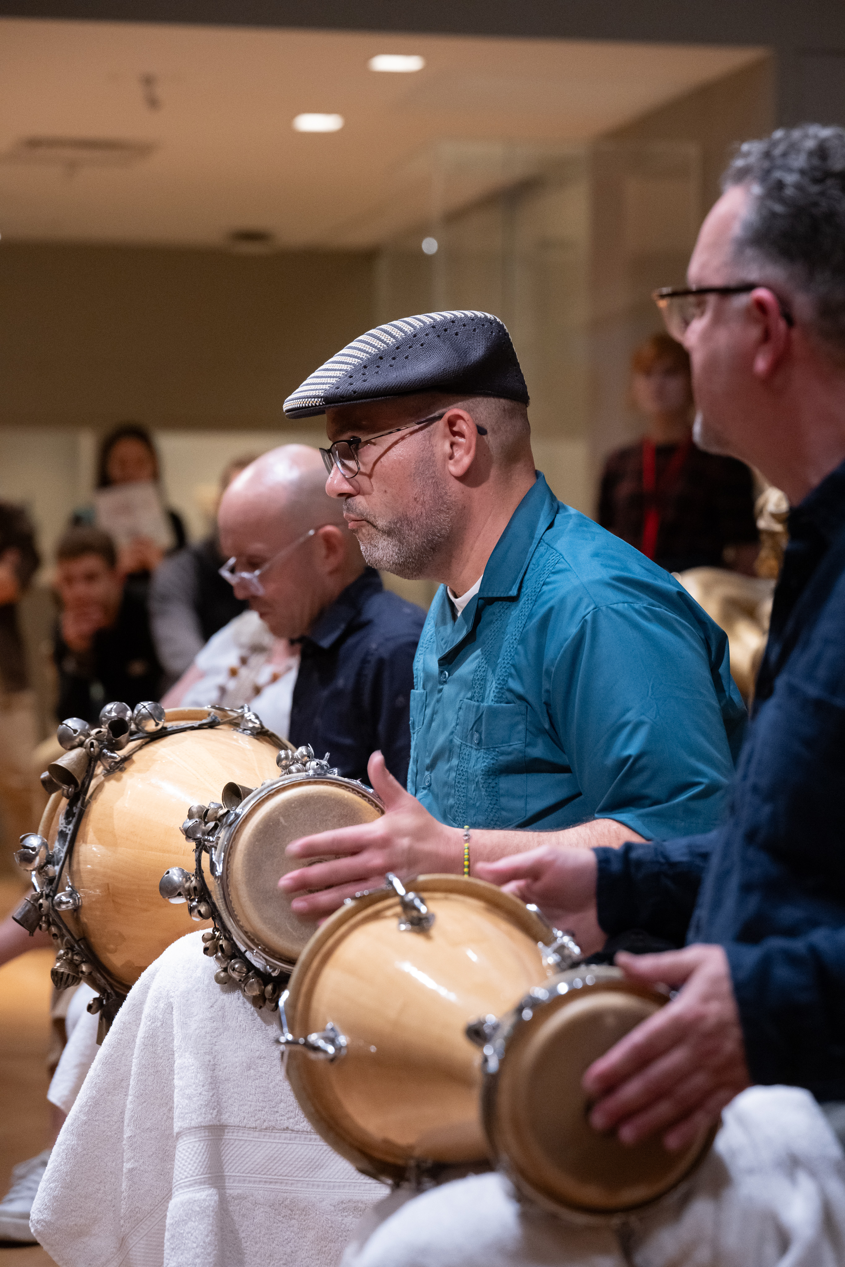A man in a blue shirt and a black hat plays a drum. He is sitting in between 2 other men who are also playing drums. 